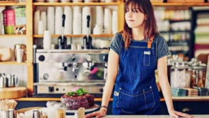 Getty Images A young woman with long reddish-brown hair and wearing a dark blue apron stands behind the counter in a brightly-lit cafe, next to an ornate chocolate cake. There is a large coffee machine on the back wall, with piles of empty take-away cups and several boxes of tea bags.