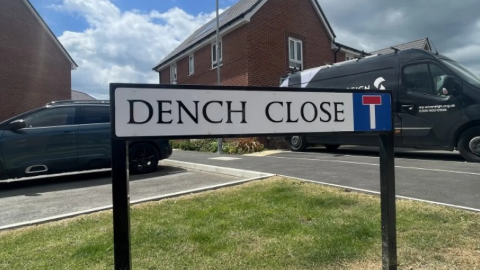 The Dench Close street sign fixed into the grass kerb side as you enter Dench Close with a van, a car and red-brick houses in the background.