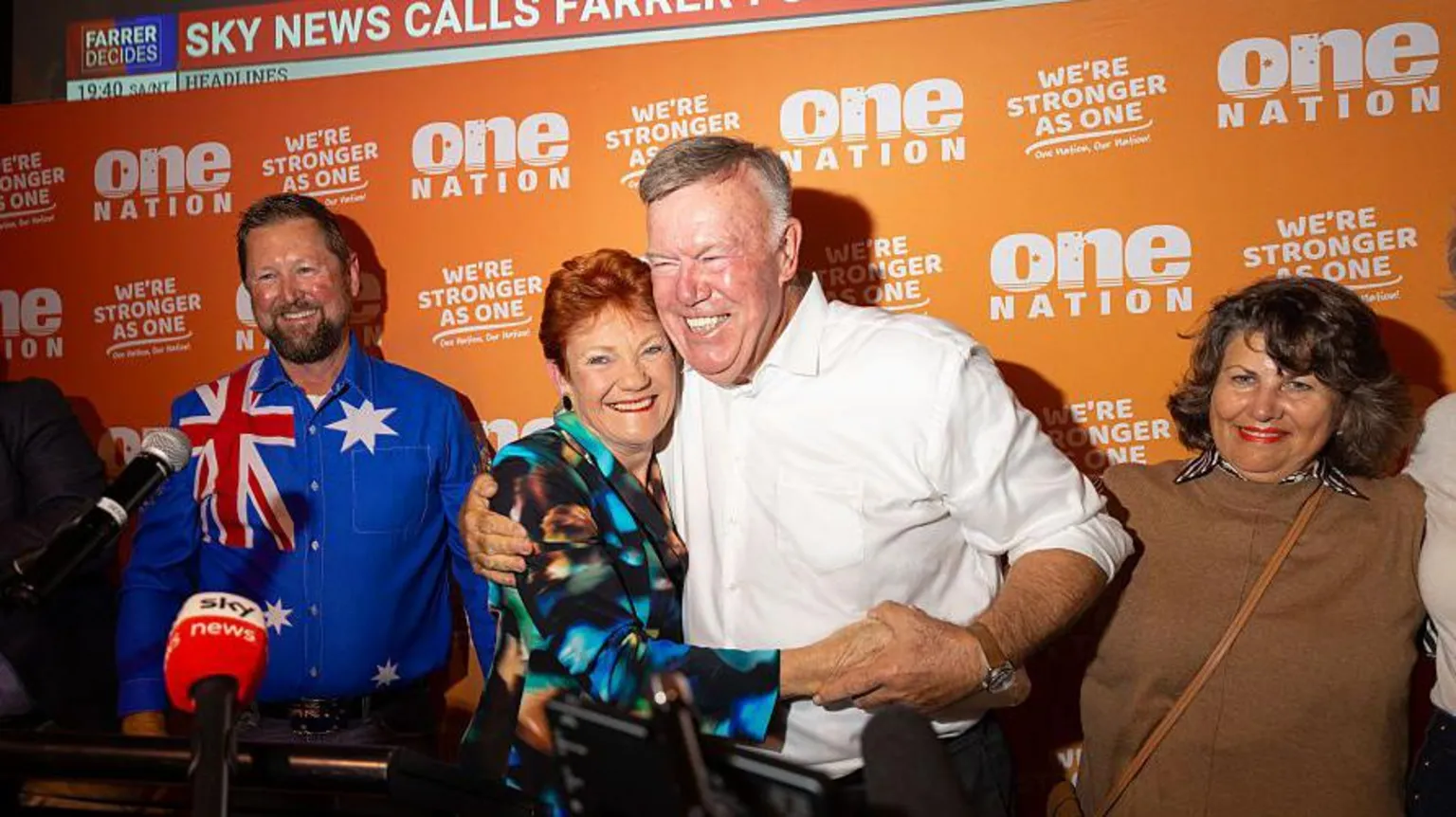 Pauline Hanson wearing a blue shirt hugs David Farley wearing a white shirt with a One Nation sign behind