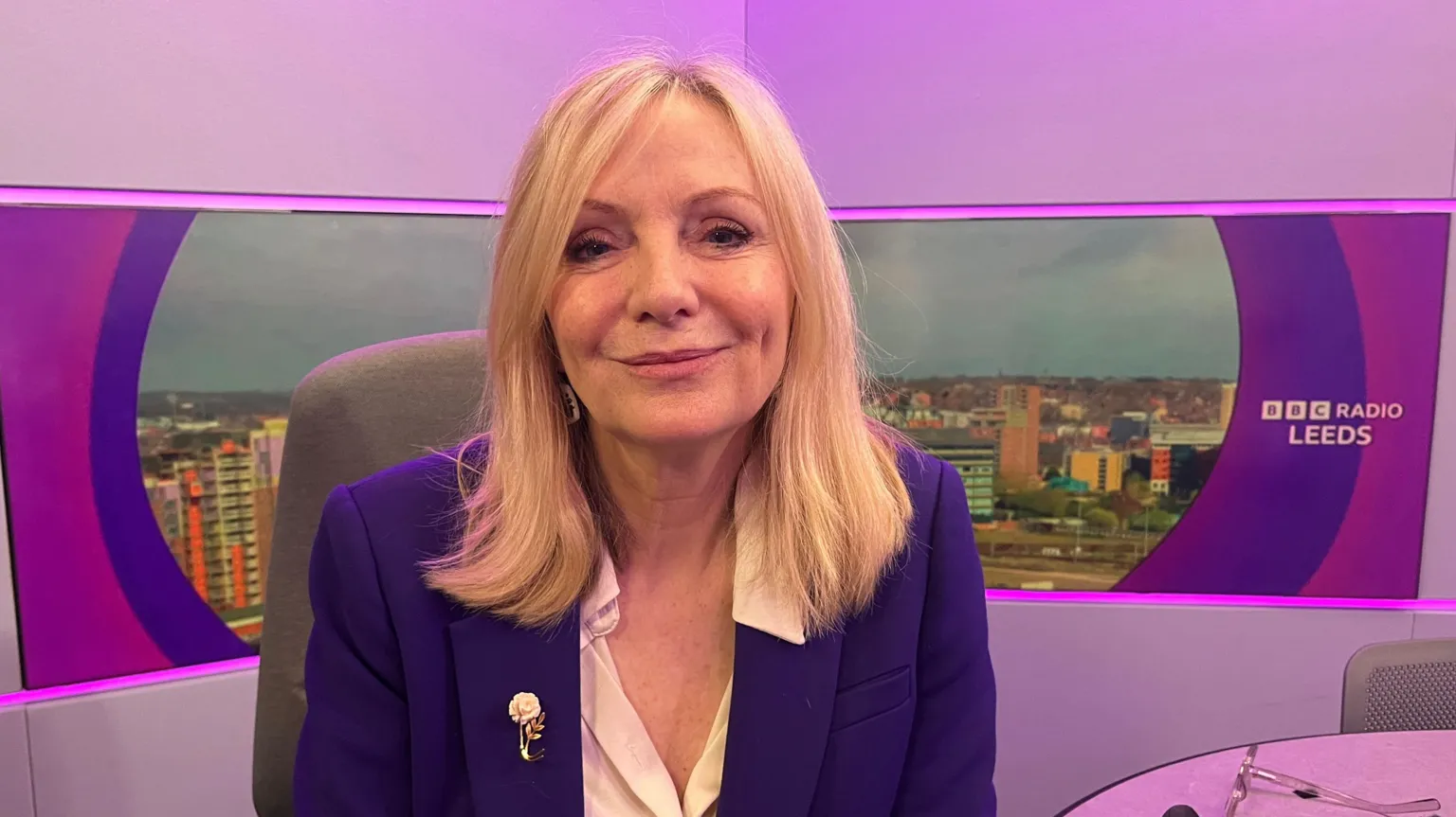 A woman with blonde hair, sitting in a radio studio. She is wearing a blue jacket and white shirt. There is a brooch on her jacket. The studio is lit in purple, and there is a skyline of Leeds behind her.