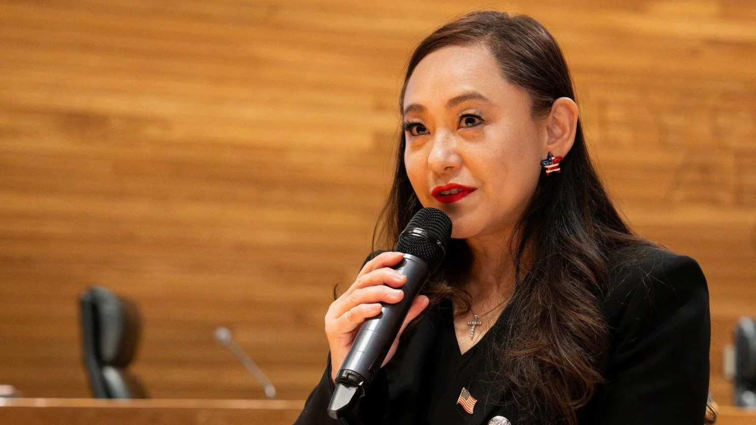 Eileen Wang, wearing a black jacket with an United States flag pin, holds a microphone as she addresses Arcadia town hall in California.
