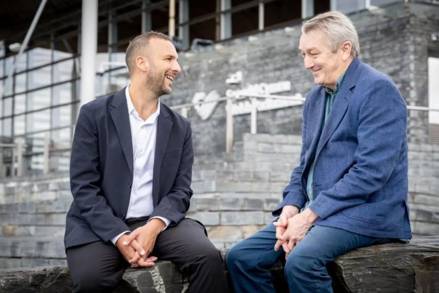 Green Party Zack Polanski and Anthony Slaughter chatting and smiling, sitting together beside the grey steps to the Senedd building in Cardiff Bay.