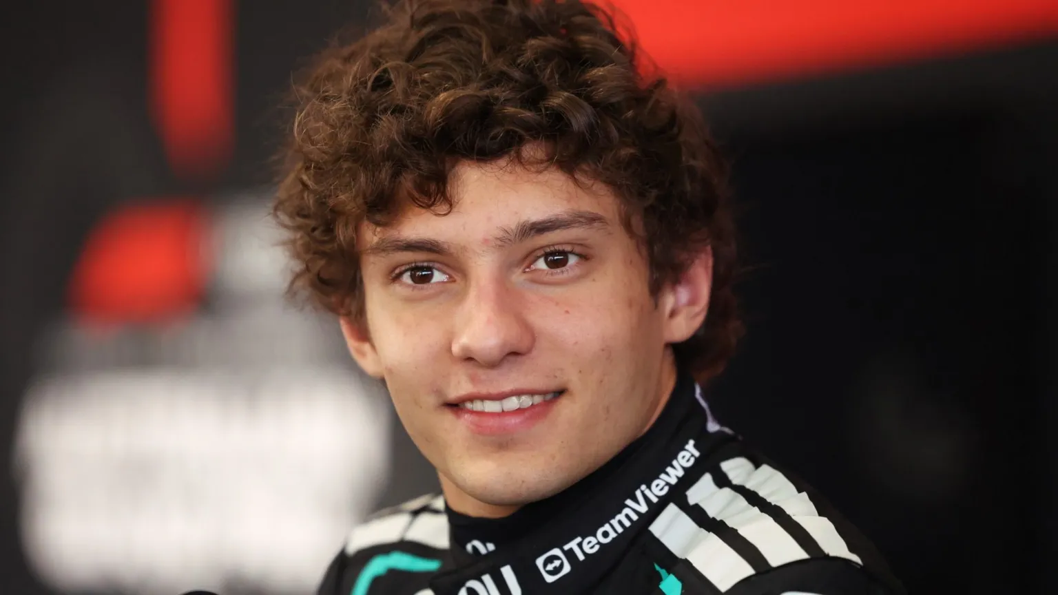 A young motorsport driver with curly brown hair, dressed in a sleek black racing suit accented with white and teal details. Sponsor logos are visible across the suit, including one on the collar.