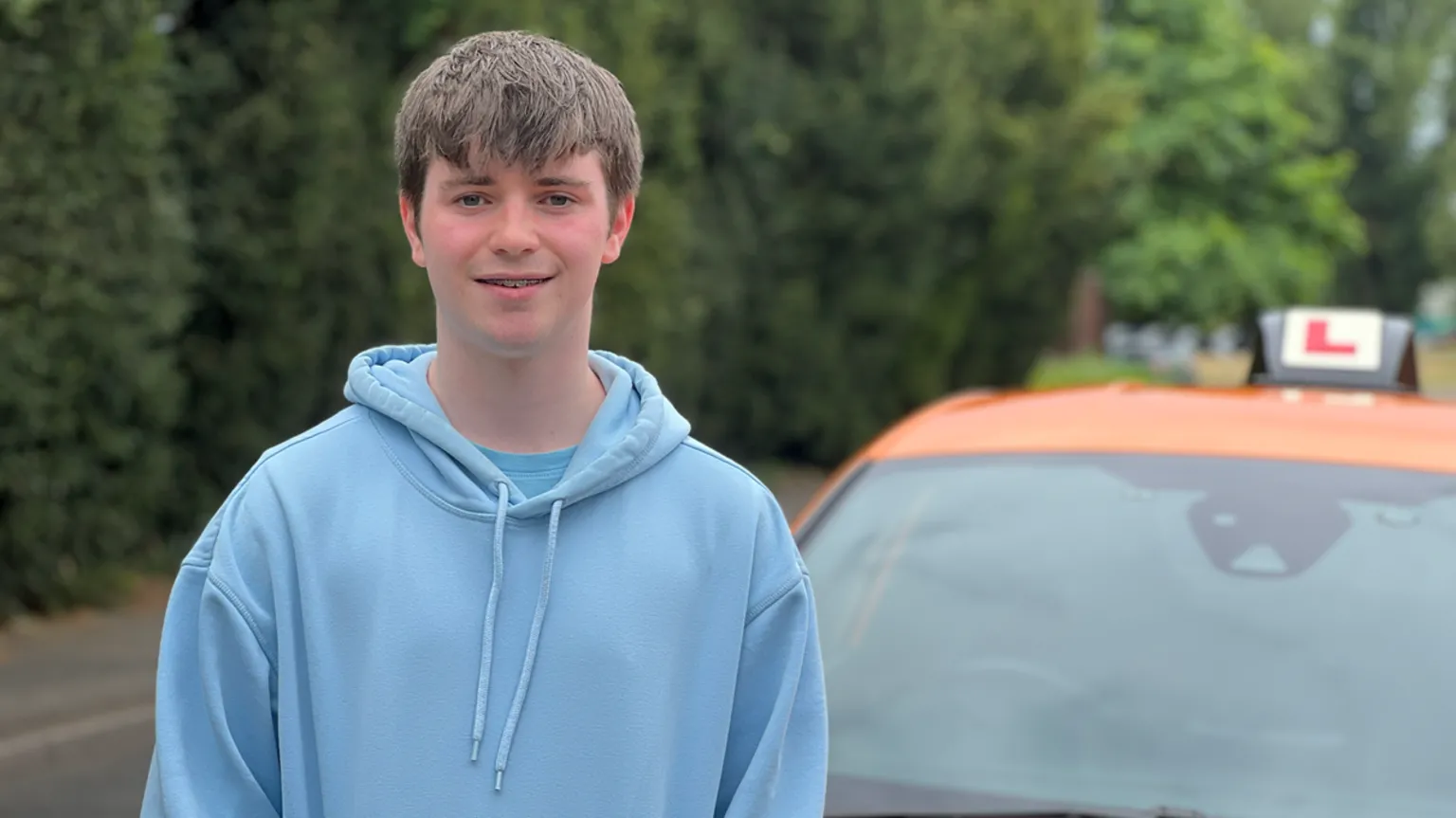 Alex has straight, brown hair and wears a light blue hoodie. He stands next to a driving school car.