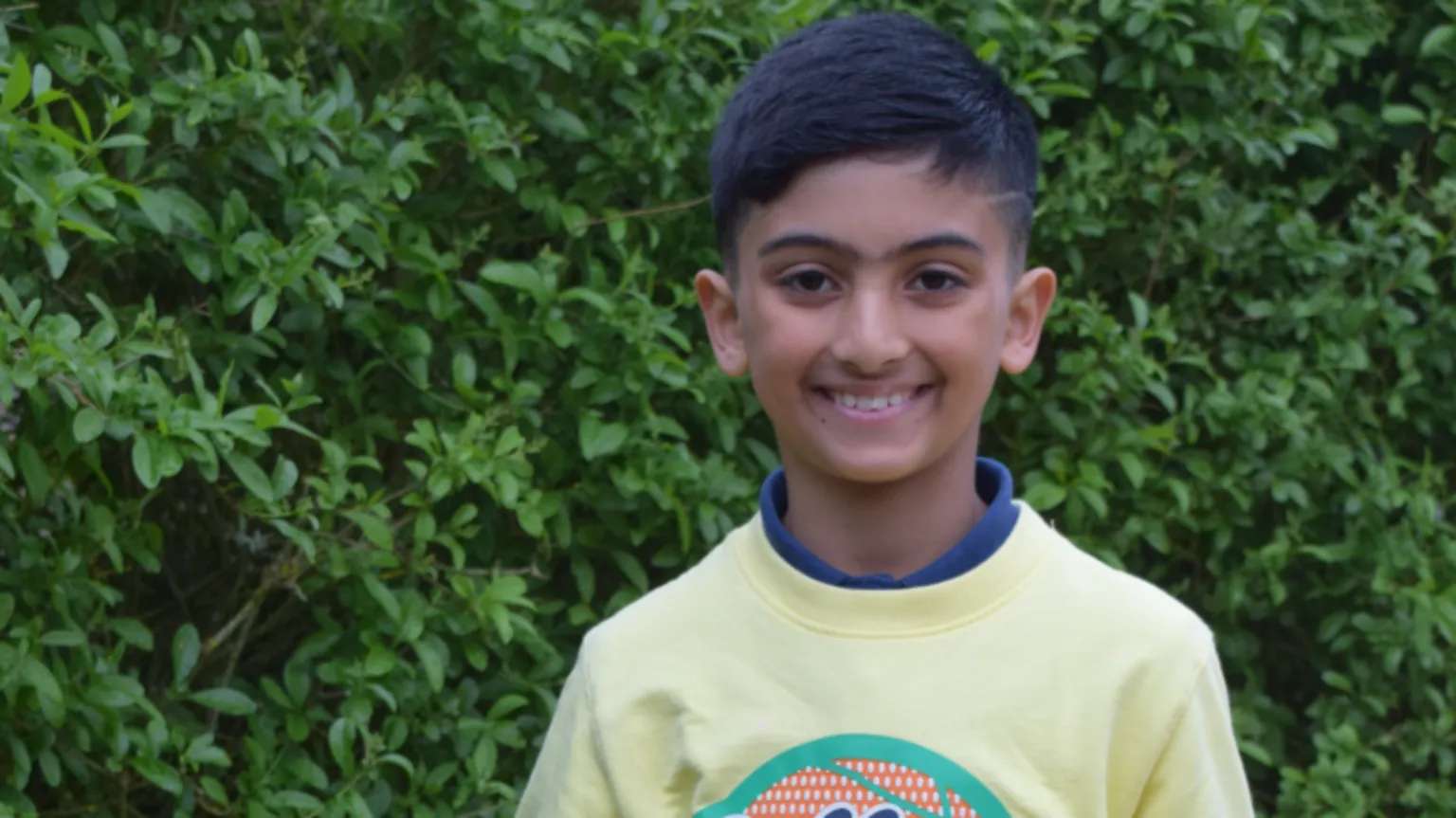 Kayaan has dark hair and eyes and is smiling at the camera. He is wearing a yellow jumper and is standing in front of a hedge.