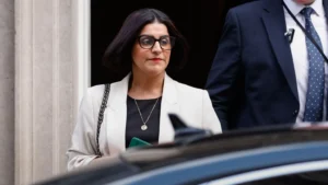 EPA Home Secretary Shabana Mahmood walking to a car wearing dark brown glasses, red lipstick, and a light grey blazer.