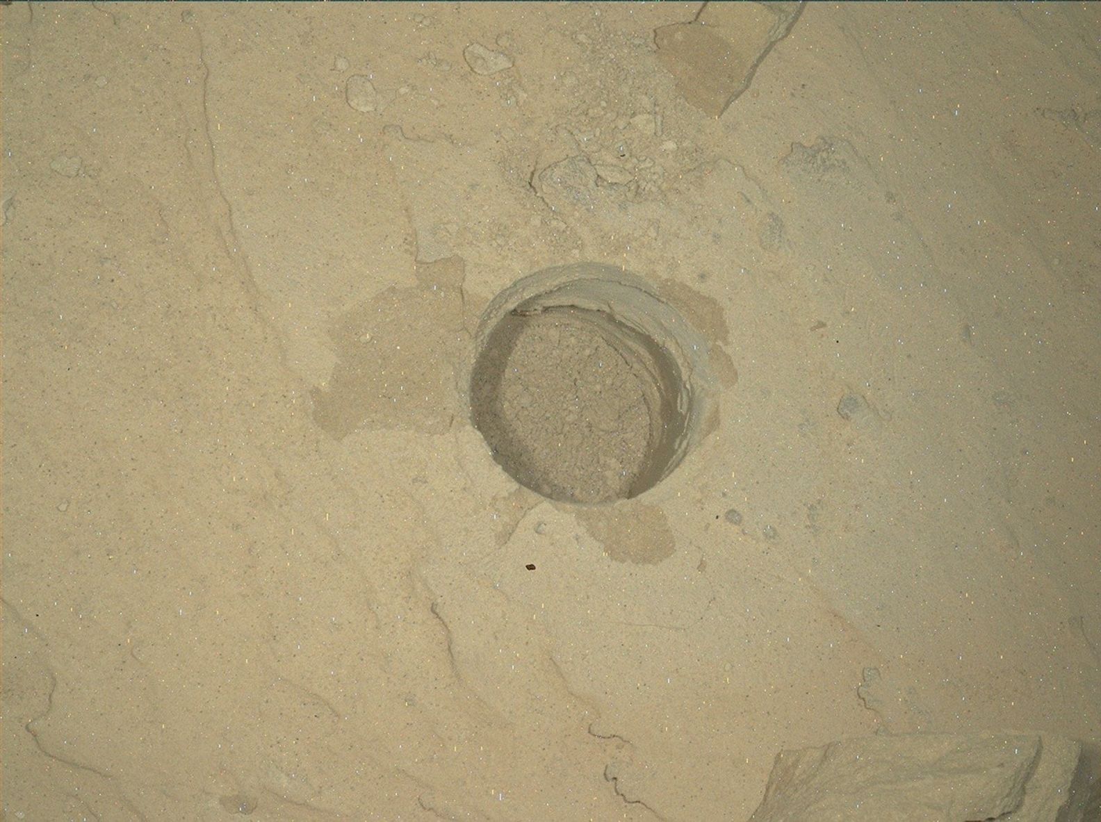 A close-up view of a perfectly circular drill hole in light tan Martian bedrock, created by the Curiosity rover. The shallow hole is filled with loose, pale, powdery rock cuttings. The surrounding rock surface is mostly flat and dusty, featuring subtle ridges, faint cracks, and a few small, scattered pebbles.