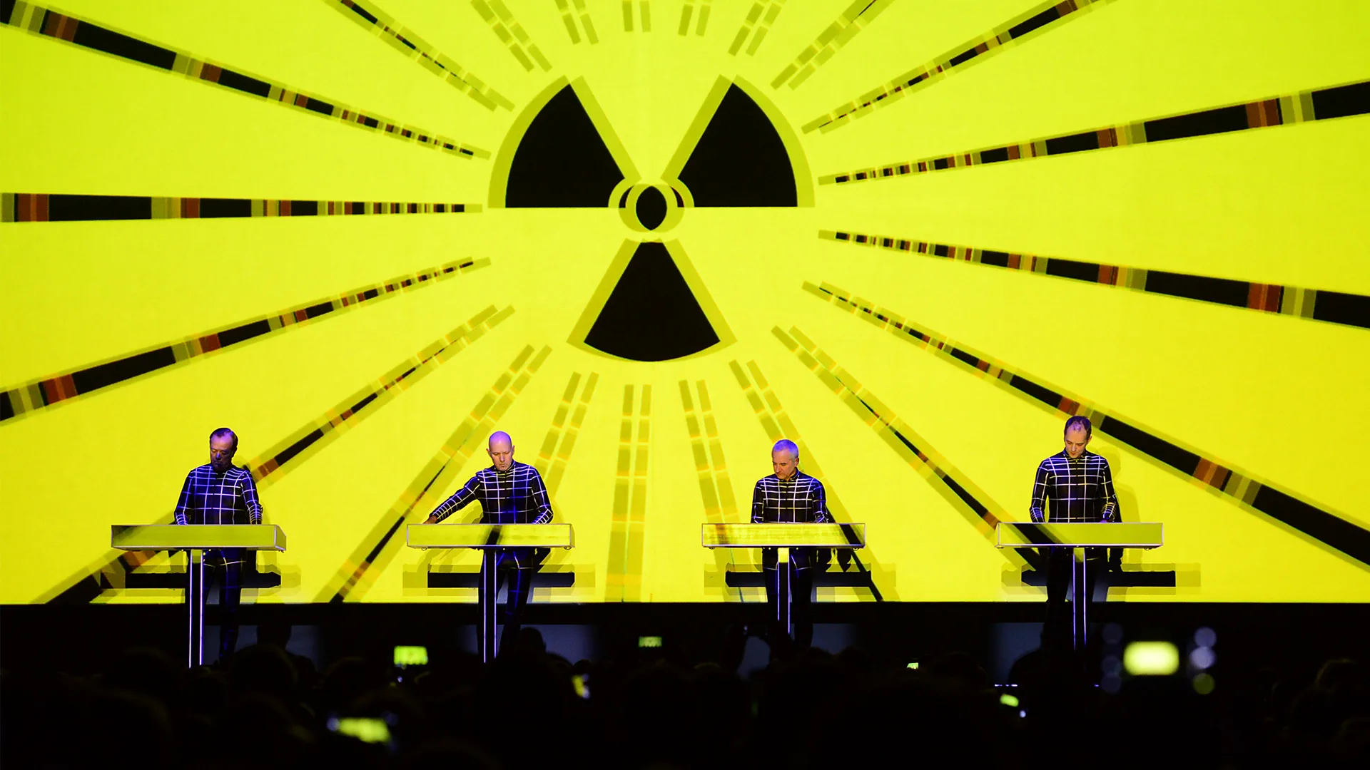 The four members of Kraftwerk performing on stage with a radioactive symbol against a bright neon yellow screen behind them (Credit: Getty Images)